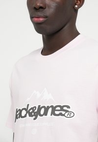 Ροζ βαμβακερό T-shirt με το λογότυπο "jack&jones" σε μαύρο, �με λευκά γραφικά ορεινών τοπίων από πάνω και το κείμενο "STANDARD ISSUE SUPPLIES" από κάτω.