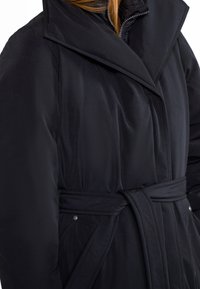 Vrouw in een zwarte winterjas met ceintuur en brede kraag, met een uitritsbare binnenlaag, getoond van midden torso tot nek.