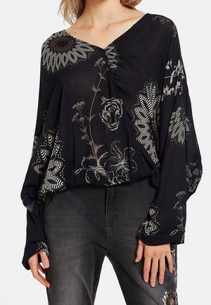 Mujer con blusa negra de manga larga con estampados blancos de flores y cara de tigre, combinada con vaqueros gris oscuro con bordados en el muslo.