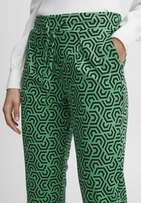 Grüne gemusterte Hose mit einem geometrischen schwarzen Design, elastischem Bund und Seitentaschen. Das Material wirkt glatt mit einem leichten Glanz.