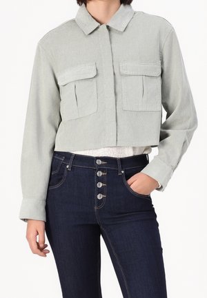 Veste courte grise avec poches à rabat portée sur un top blanc, associée à un jean bleu foncé taille haute avec quatre boutons à l'avant.