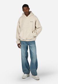 Κρέμα χρώματος φλις φούτερ με κουκούλα, μπροστινές τσέπες και ελαστικούς καρπούς, σε συνδυασμό με ανοιχτόχρωμα παντελόνια με φαρδιά πανιά και λευκά αθλητικά παπούτσια.