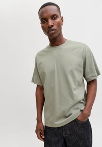 Junger Mann mit kurzen Haaren, der ein hellgrünes T-Shirt und dunkle Jeans trägt und mit einer Hand in der Tasche vor einem einfarbigen hellen Hintergrund steht.