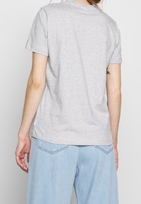 Persona vestida con una camiseta de manga corta gris claro y pantalones de mezclilla azul claro, vista desde atrás contra un fondo liso.