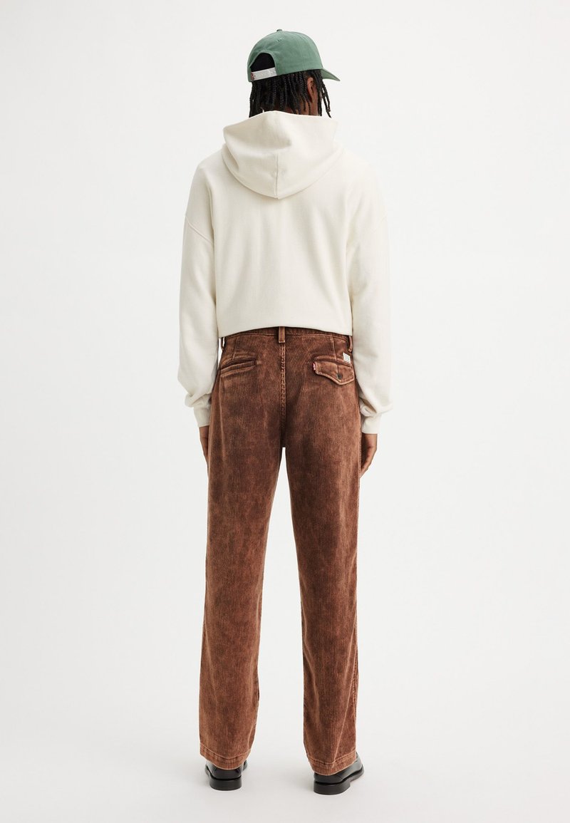 Braune Cordhose mit weitem Bein, ausgestattet mit zwei Gesäßtaschen, kombiniert mit einem weißen cropped Hoodie und einer grünen Mütze.