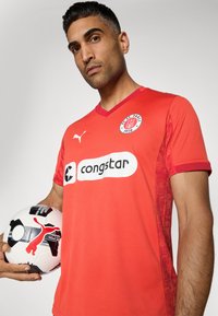 Maillot de football rouge avec col en V et logo du FC St. Pauli. Présente un motif texturé sur les côtés et un logo de sponsor blanc. Tient un ballon noir et blanc.