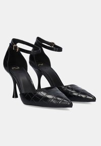 Zapatos de tacón alto de charol negro con un patrón acolchado, punteras afiladas, correas en los tobillos y tacones delgados.