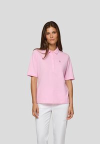 Helles rosa Polohemd mit kurzen Ärmeln, Knopfleiste und Logo auf der linken Brust. Kombiniert mit weißen, ausgewaschenen Jeans.