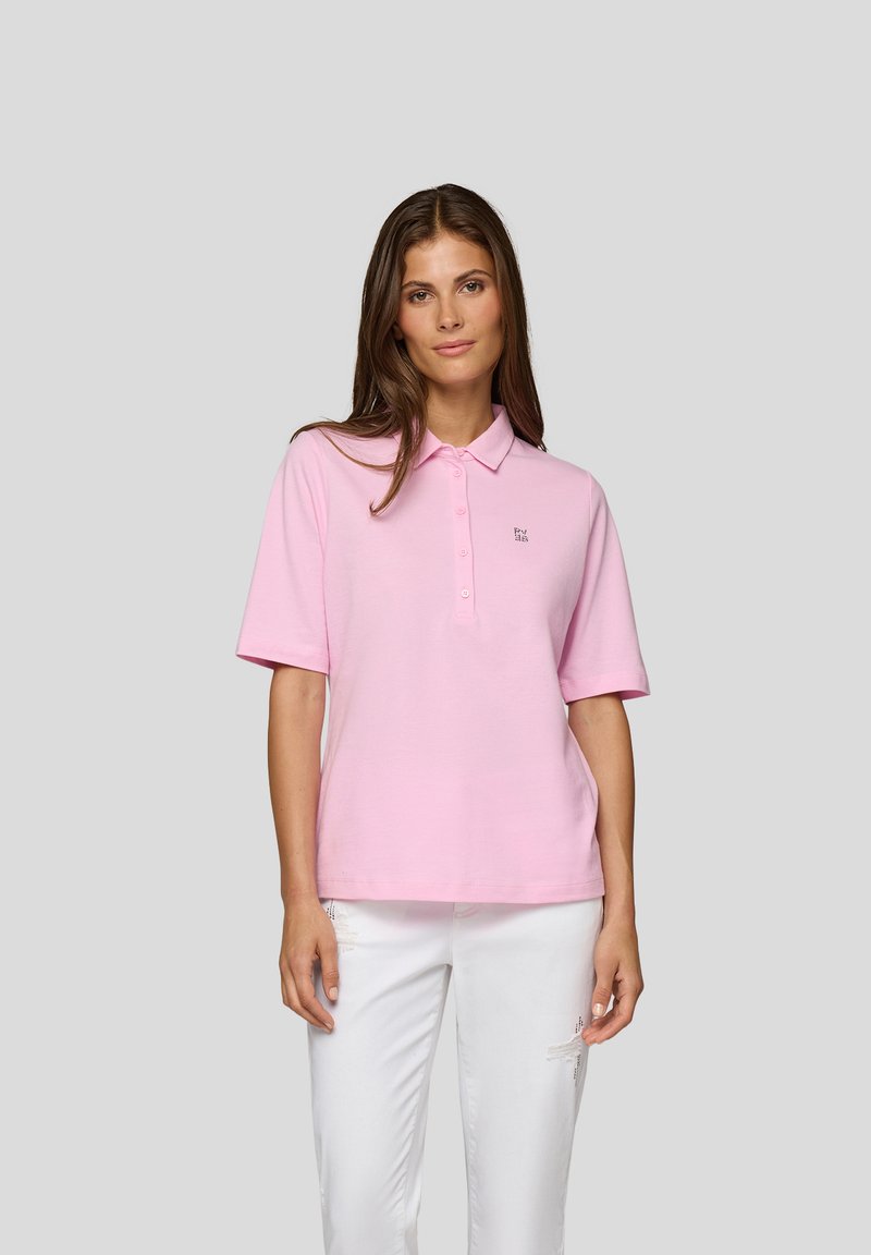 Helles rosa Polohemd mit kurzen Ärmeln, Knopfleiste und Logo auf der linken Brust. Kombiniert mit weißen, ausgewaschenen Jeans.