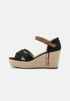 Sandalia de cuña con tiras de tela negra, hebilla marrón en el tobillo y plataforma de yute trenzado con detalles en franjas rojas, blancas y azules.