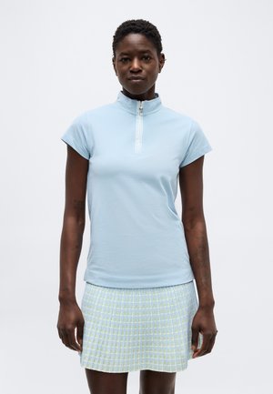 Vrouw draagt een lichtblauw topje met korte mouwen en een rits aan de voorkant en een lichtblauwe en gele geruite rok, staand tegen een witte achtergrond.