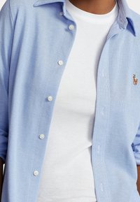 Ljusblå knapp-up skjorta med en texturerad tyg, med en vit t-shirt under och en liten broderad logotyp på bröstet.