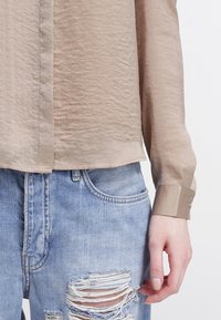 Gros plan d'une personne portant une chemise beige à manches longues et un jean bleu clair déchiré, avec une main posée près de la zone déchirée.