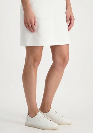 Kvinde iført en hvid nederdel og hvide lave snøresneakers står mod en ensfarvet baggrund.