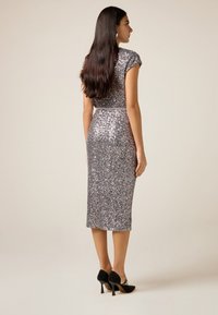 Kvinde med langt mørkt hår iført en sølvglitrende midikjole og sorte højhælede sko, stående i profil mod en ensfarvet baggrund.