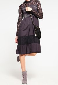 Vestido morado oscuro con mangas de encaje y bordado floral, que presenta una banda central texturizada y un dobladillo con forma de concha. Combinado con botas de tobillo grises.