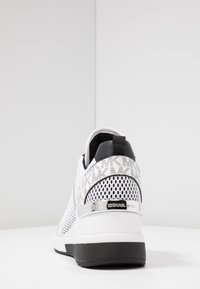 Sneaker bianca in mesh con accenti neri, lati texturizzati e un dettaglio del logo metallico sul tallone. Presenta un collare imbottito e una suola bianca.