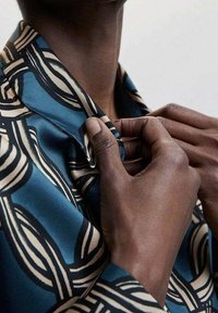 Chemise en soie sur fond bleu, présentant un motif abstrait noir et beige. Boutons au col, mains ajustant le tissu.