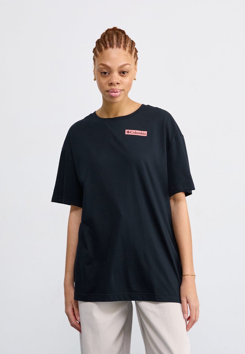 Camiseta de algodón negra con cuello redondo, mangas cortas y un parche de logo rosa en el lado izquierdo. Llevada con pantalones de color claro.