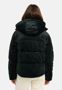 Veste noire en style doudoune avec une grande capuche, finition texturée et détails de couture visibles. Le tissu semble doux et moelleux.