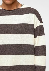 Gestreepte trui in bruin en crème kleuren, van textuur gebreid materiaal, brede strepen en een rauwe halslijn. Mouwen zijn iets oversized.