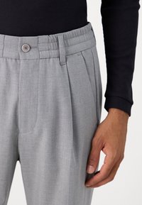 Pantalons gris clair avec une texture lisse, devant plissé, poches angulaires et une fermeture à un bouton au niveau de la taille.