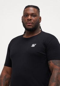 Tricou negru cu mânecă scurtă, fabricat din bumbac, având un guler rotund și un mic logo alb pe partea stângă a pieptului.