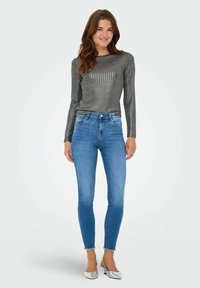 Metaalgrijze langemouwtop met verticale ribbels, gecombineerd met strakke blauwe jeans met een gerafelde zoom en versierde zakken. Zilveren flats.