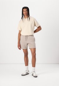 Camiseta de algodón beige combinada con pantalones cortos de cuerda de color gris claro. Se lleva con calcetines blancos y zapatillas deportivas con acentos en negro y gris.