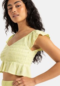 Light yellow cropped top with a smocked texture, ruffled sleeves, and a peplum hem. Fabric appears soft and lightweight. Small hardware detail visible.