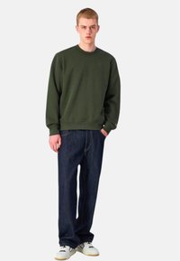 Felpa verde scuro con scollo tondo, abbinata a jeans blu scuro a vita alta e scarpe da ginnastica bianche. Design semplice con minimalismi.