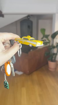 Žltý model hračkárskeho taxíka držený nad súborom kľúčov, vybavený taxíkovou značkou na streche, pruhovanými nálepkami a hladkým plastovým povrchom.