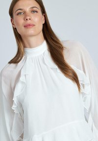 Camicia bianca con maniche lunghe e fluttuanti, caratterizzata da dettagli arricciati lungo il colletto e il corpo. Realizzata in materiale leggero e texturizzato.