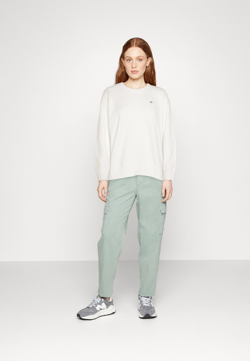 Biała bluza z okrągłym dekoltem, zestawiona z jas green spodniami cargo z bocznymi kieszeniami. Model nosi szare sneakersy. Proste tło.