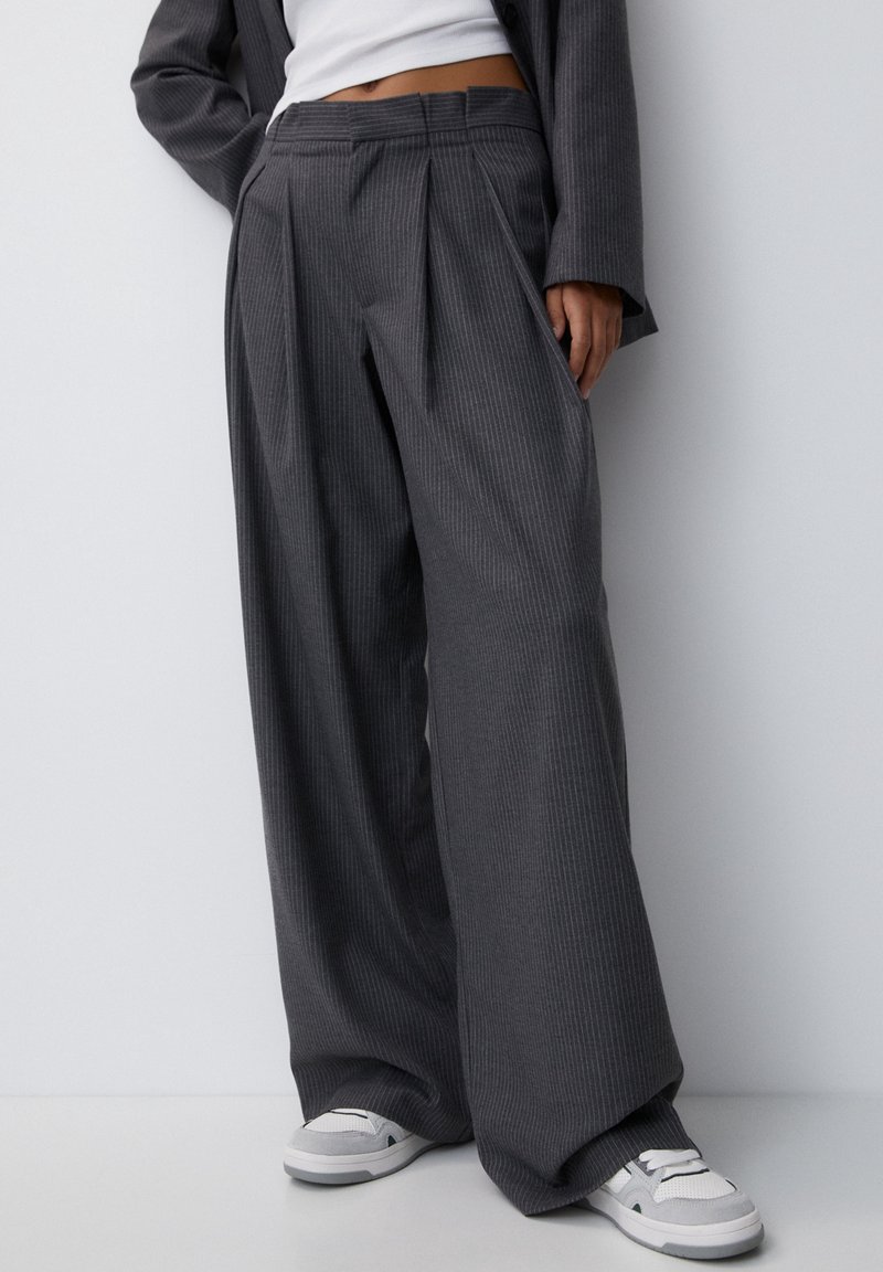 Personne portant un pantalon large gris foncé à fines rayures, un crop top blanc et des baskets blanches, debout contre un mur blanc.