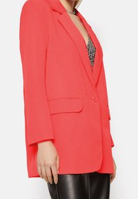 Blazer corallo con un design sartoriale, revers a scollo, bottone singolo e due tasche frontali, abbinato a pantaloni in ecopelle neri.