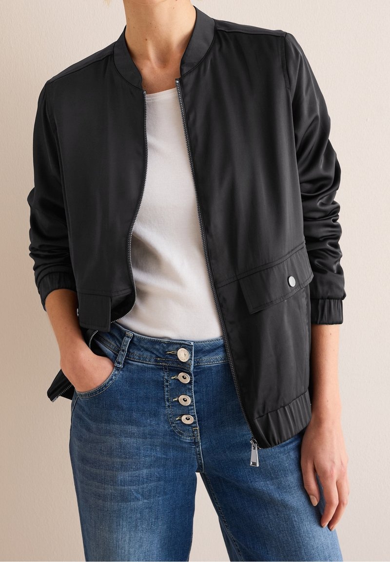Veste bomber noire avec une texture lisse, fermeture éclair à l'avant et poignets élastiques, associée à un jean bleu clair avec un design à cinq boutons.