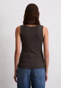 Frau mit schulterlangem braunem Haar, die ein ärmelloses dunkelbraunes Oberteil und blaue Jeans trägt, vor einer schlichten hellen Wand stehend.