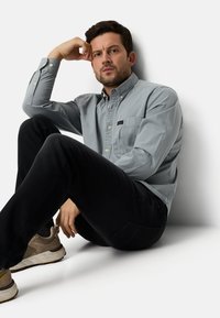 Hellgraues Hemd mit Knopfleiste und Brusttasche, kombiniert mit schwarzer Jeans. Das Modell sitzt lässig an einer Wand und trägt beigefarbene Sneakers.