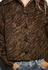 Veste en pied-de-poule marron avec un col, dotée de deux rangées de boutons noirs et d'un tissu texturé. Portée sur un jean gris.