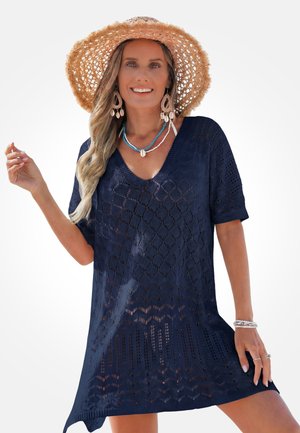 Woman smiling while wearing a navy blue patterned knit dress, wide-brim straw hat, shell earrings, turquoise necklace, and silver bracelets.