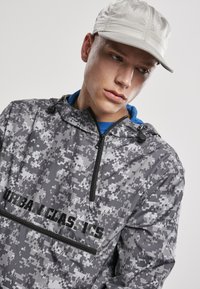 Jachetă pulover de camuflaj gri cu fermoar pe față, glugă cu șnur și logo-ul "URBAN CLASSICS" în negru. Purtați cu o șapcă gri deschis.