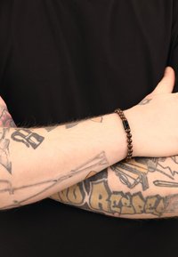 Brązowa bransoletka z koralikami z czarnym akcentem, nawleczona na elastyczną nitkę, noszona na przedramieniu pokrytym tatuażami, na czarnym tle.