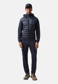 Mørkeblå zip-up polstret vest over en mørkeblå hoodie, kombineret med matchende sweatpants og brune/grå sneakers. Accentueret med en mørk baseballkasket.