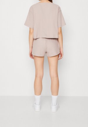 Vrouw staand met haar rug naar de camera, gekleed in een beige shirt met korte mouwen en bijpassende korte broek, witte sokken en witte sneakers op een effen achtergrond.