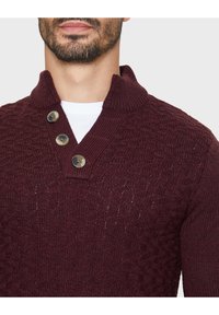 Pullover in magenta lavorato a maglia con un motivo testurizzato, caratterizzato da un collo a scialle, tre bottoni e una vestibilità aderente. Camicia bianca sotto.