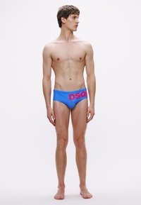 Sinised ujumispüksid, millel on kontrastne roosa "DSQ" logo, sile tekstuur, keha järgnev lõige ja keskmise kõrgusega vöökoht, mudel seisab ühevärvilise tausta ees.