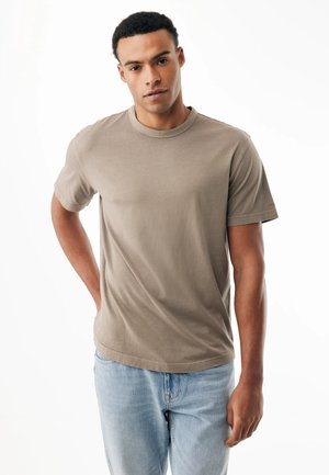 Junger Mann mit lockigem Haar, der ein schlichtes beiges T-Shirt und hellblaue Jeans trägt, mit einer Hand in der Tasche vor weißem Hintergrund stehend.