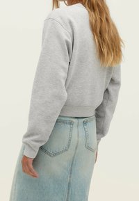 Une personne avec de longs cheveux blonds portant un sweat-shirt gris clair et une jupe en denim bleu clair taille haute, vue de dos.