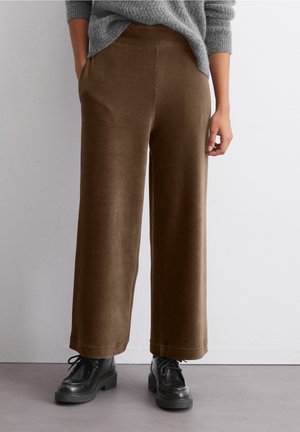 Personne portant un pantalon large en velours côtelé marron, un pull gris et des chaussures noires à lacets, debout contre un mur clair uni.